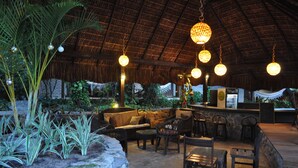 Un espacio de asientos al aire libre con techo de paja, mobiliario de mimbre e iluminación esférica colgante.