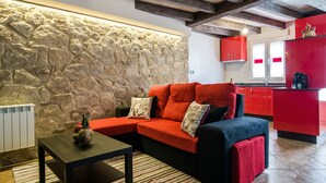 A cozy living room with a stone wall, a red and blue sofa, a small table with a vase, and a kitchen in the background. - Casa Carla (Palas de Rei)