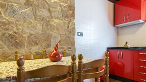 A dining area with a stone wall, a wooden table with a floral tablecloth, and red cabinets. - Casa Carla (Palas de Rei)