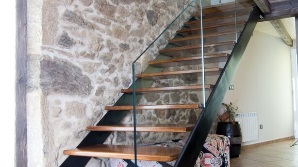 A wooden staircase with glass railings, leading to a room with a stone wall and a tiled floor. - Casa Carla (Palas de Rei)