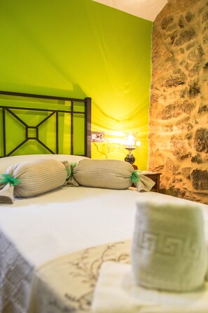 A bed with striped pillows and a white towel, a nightstand with a lamp, and a stone wall. - Casa Carla (Palas de Rei)