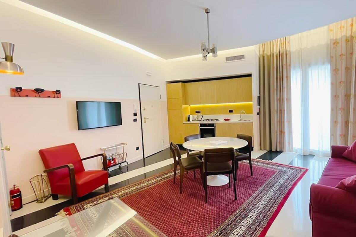 Una sala de estar moderna con un sofá rojo, una silla roja, una mesa de comedor con cuatro sillas, un televisor de pantalla plana montado en la pared y una zona de cocina con una encimera amarilla.