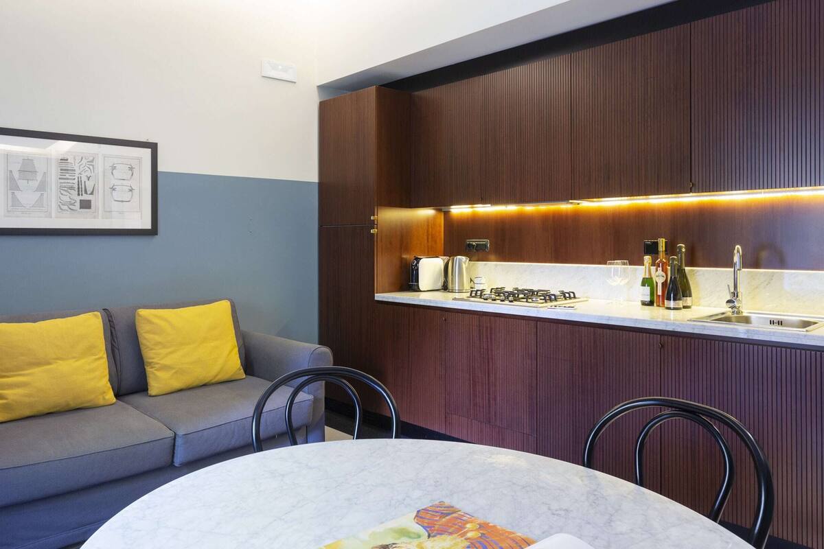 Un espacio habitable pequeño con kitchenette, un sofá con cojines amarillos, una mesa de comedor y un dibujo arquitectónico enmarcado en la pared.