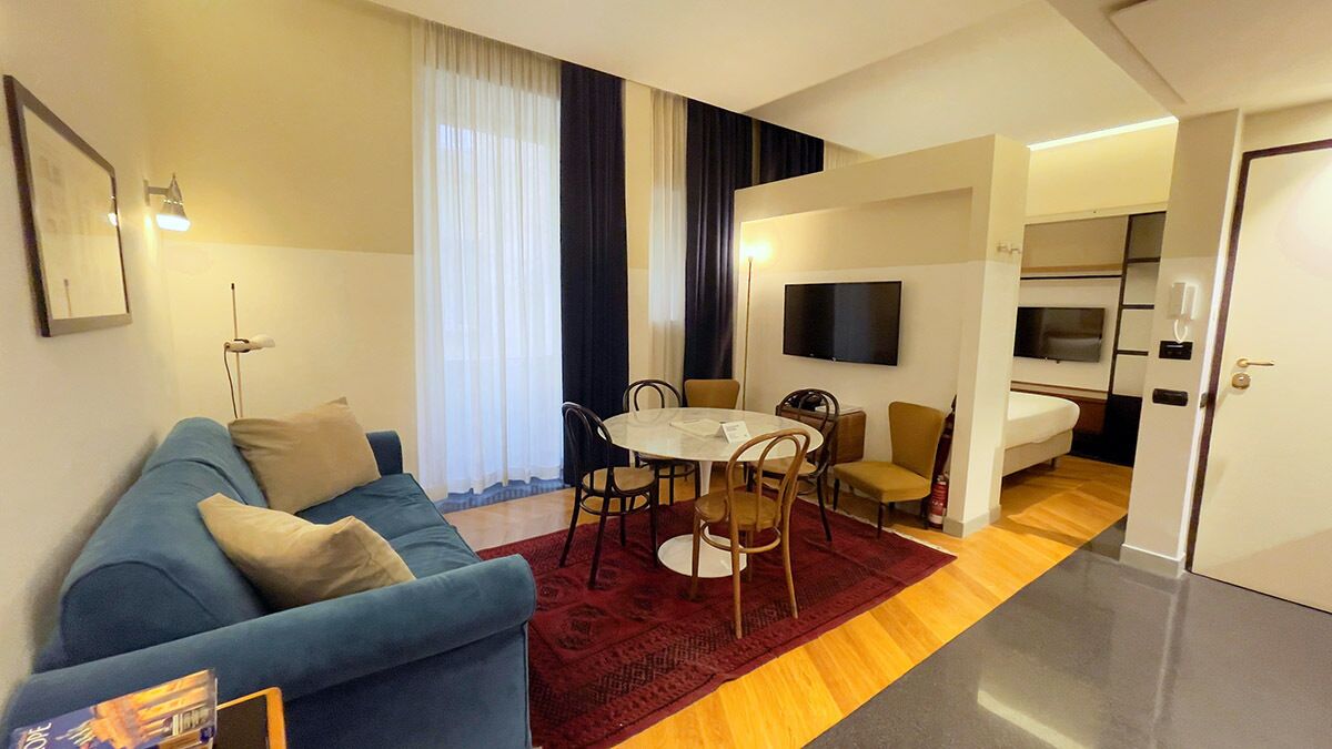 A modern hotel room with a blue sofa, a dining table with chairs, a TV mounted on the wall, and a separate kitchenette area.
