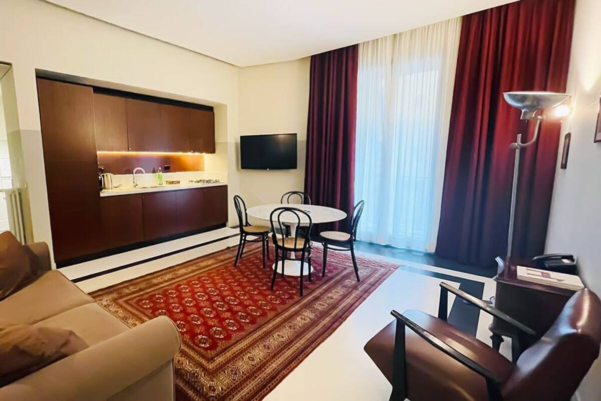 Una sala de estar moderna con sofá, una mesa de comedor con sillas, una cocina con armarios y un televisor, y un ventanal con cortinas.