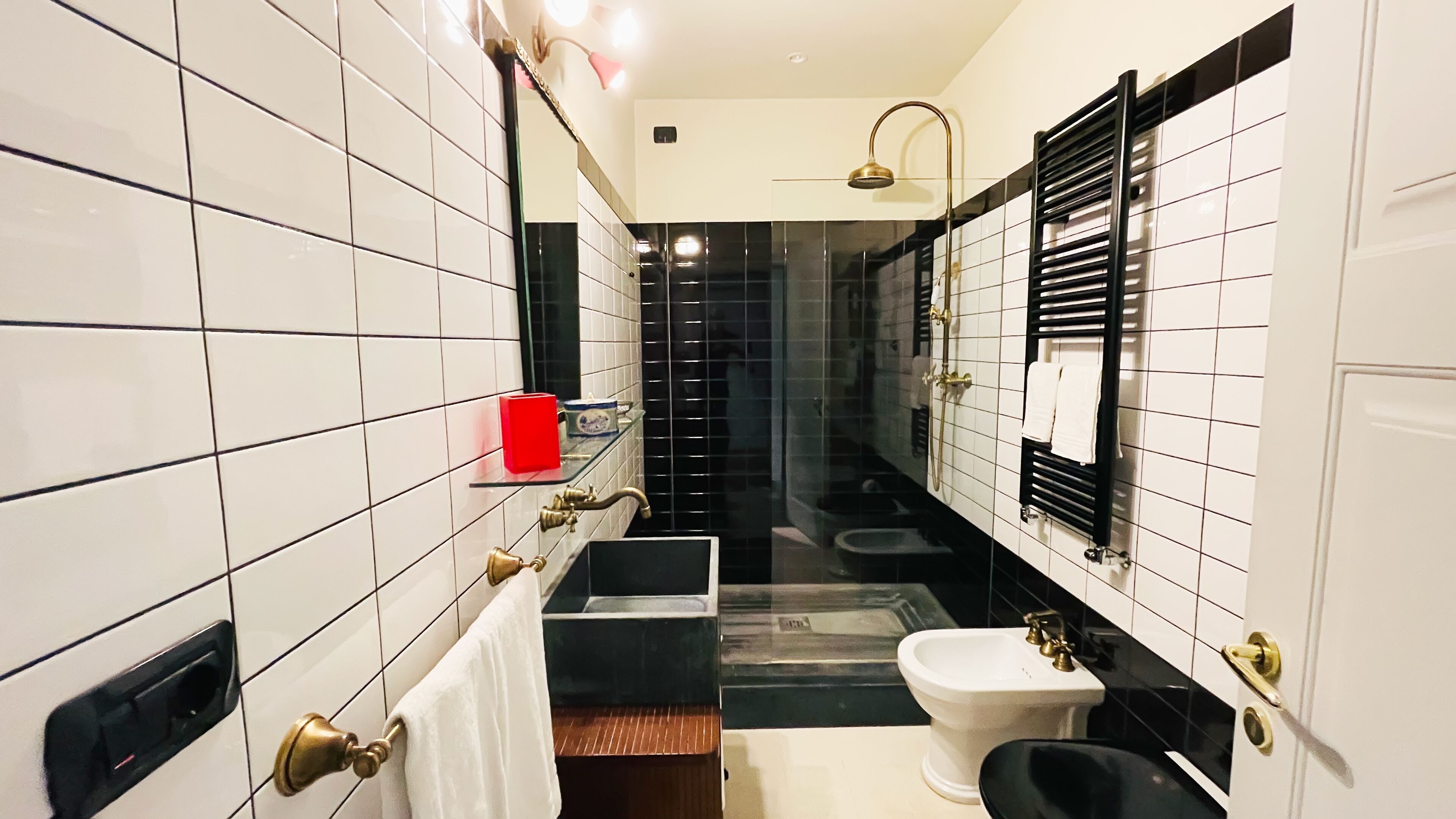 Ein schmales Badezimmer mit weiß-schwarz gefliesten Wänden, einer schwarzen Badewanne, einem weißen Waschbecken und einem roten Seifenspender.