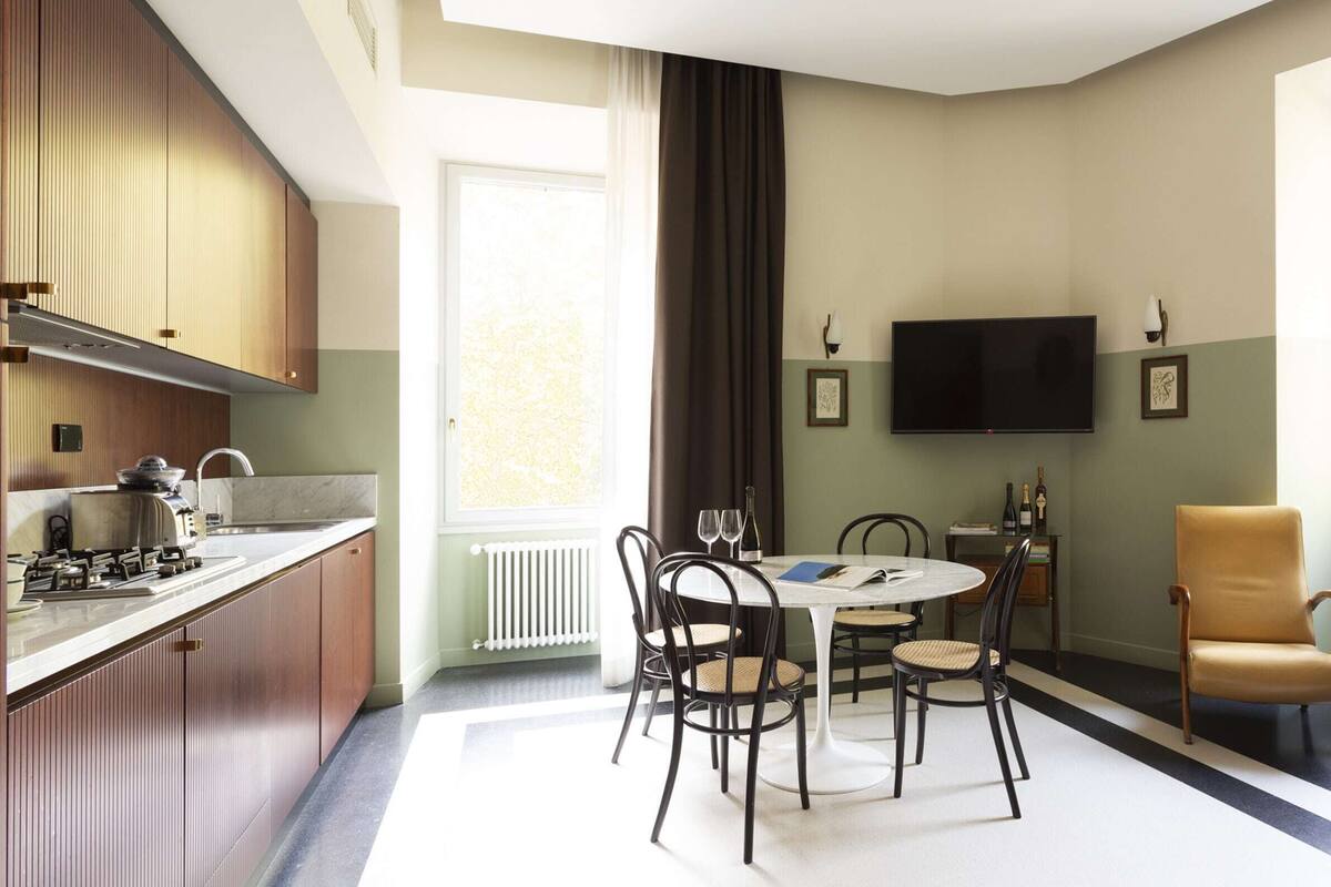 Una cocina moderna con estufa a gas, un comedor con una mesa redonda y sillas, un televisor de pantalla plana montado en la pared y una ventana con vista a la ciudad.