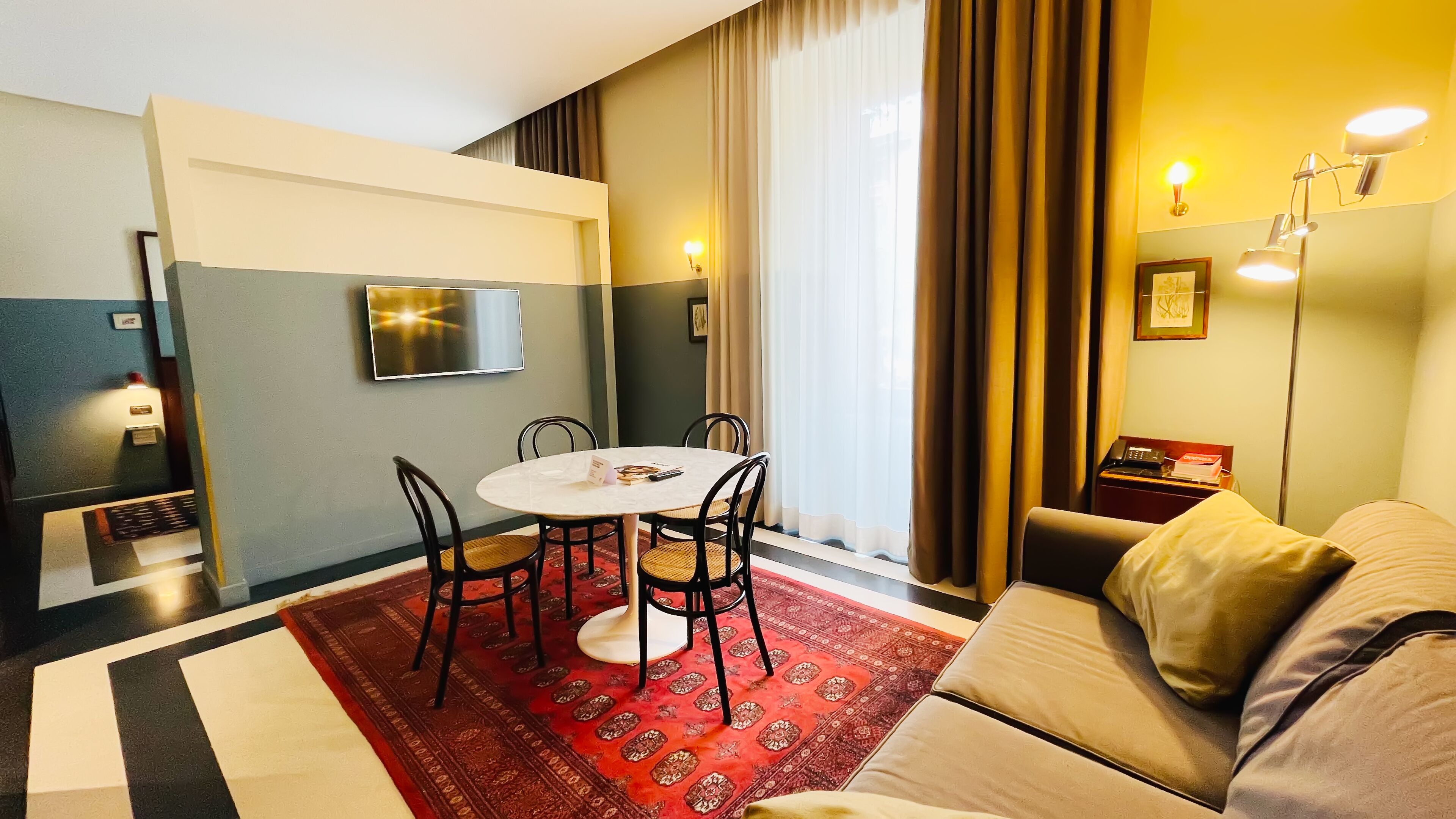 A hotel room with a round table, four chairs, a red patterned rug, a beige sofa, a TV mounted on the wall, and a bedside table with a lamp and a book.