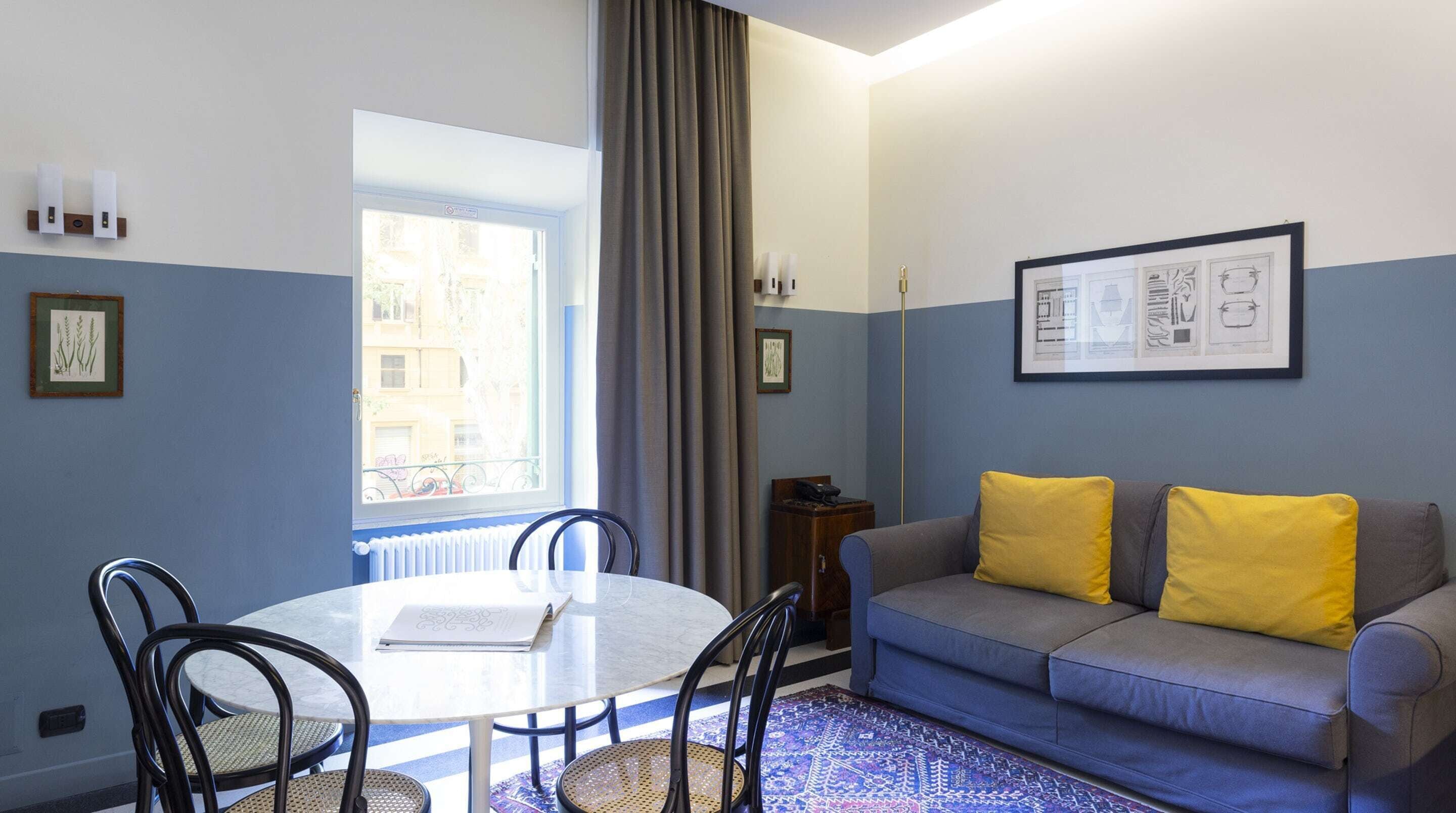 A room with a round table, two chairs, a sofa with yellow cushions, a framed picture on the wall, and a window with curtains.