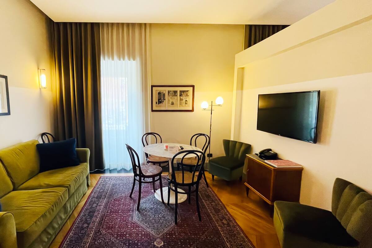 Habitación de hotel con sofá verde, una mesa de comedor con sillas, televisor montado en la pared y alfombra estampada.