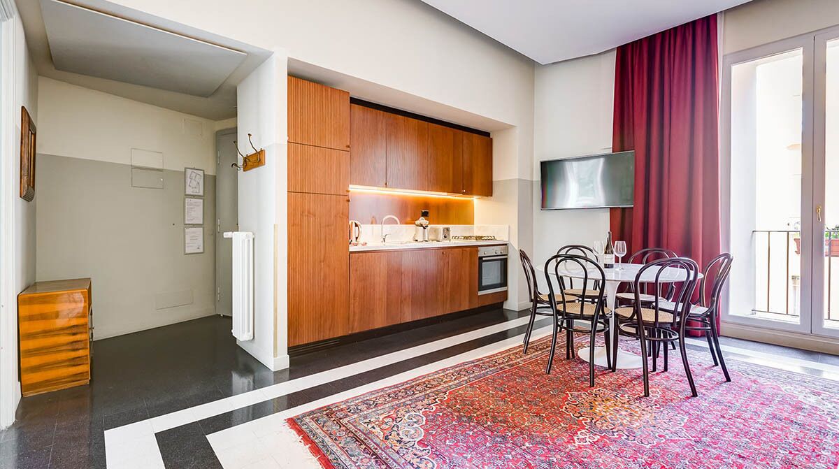 A modern kitchen with wooden cabinets, a dining area with a red rug, and a TV mounted on the wall.