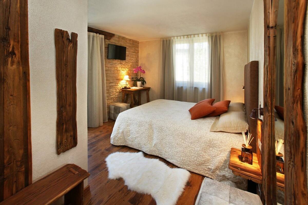 Une chambre à coucher avec un grand lit, des murs en bois, un mur d’accent en pierre, un téléviseur à écran plat et une table de chevet avec une lampe et un vase de fleurs.