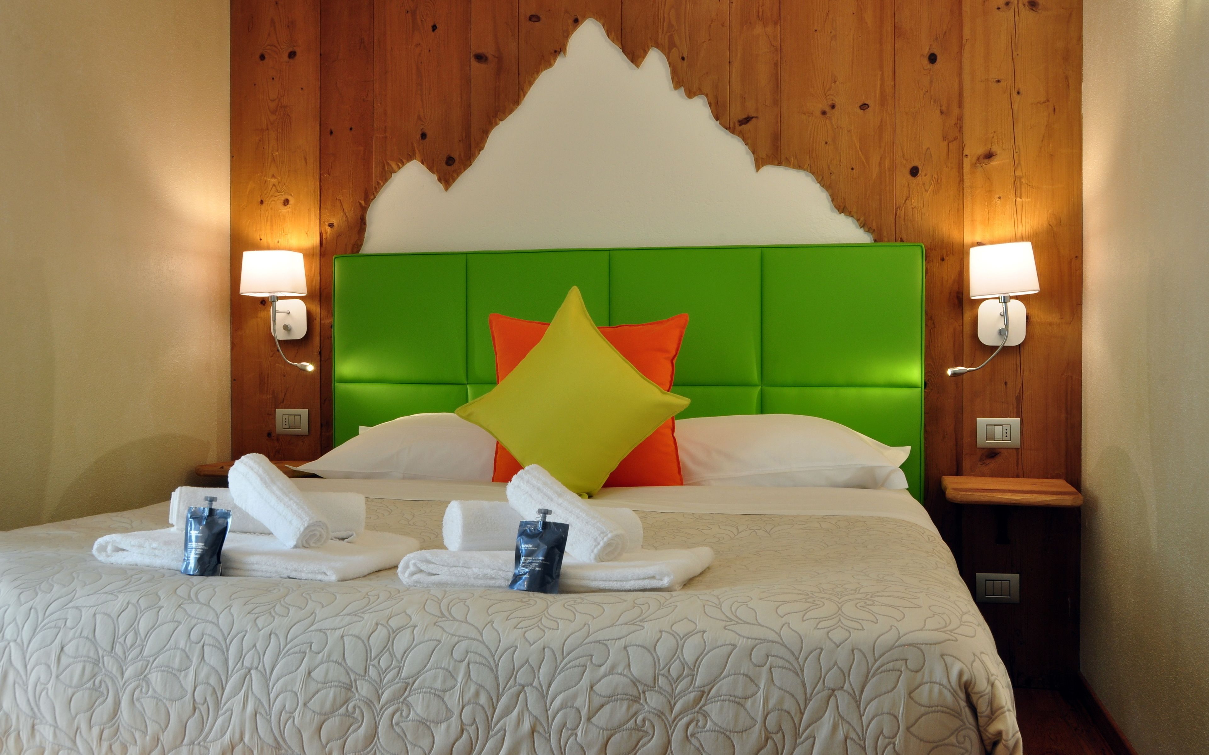 Quarto de hotel com cabeceira estofada em verde, almofadas brancas e laranja, e cama bem arrumada com toalhas e artigos de higiene.