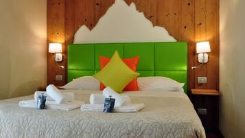 Quarto de hotel com cabeceira estofada em verde, almofadas brancas e laranja, e cama bem arrumada com toalhas e artigos de higiene.
