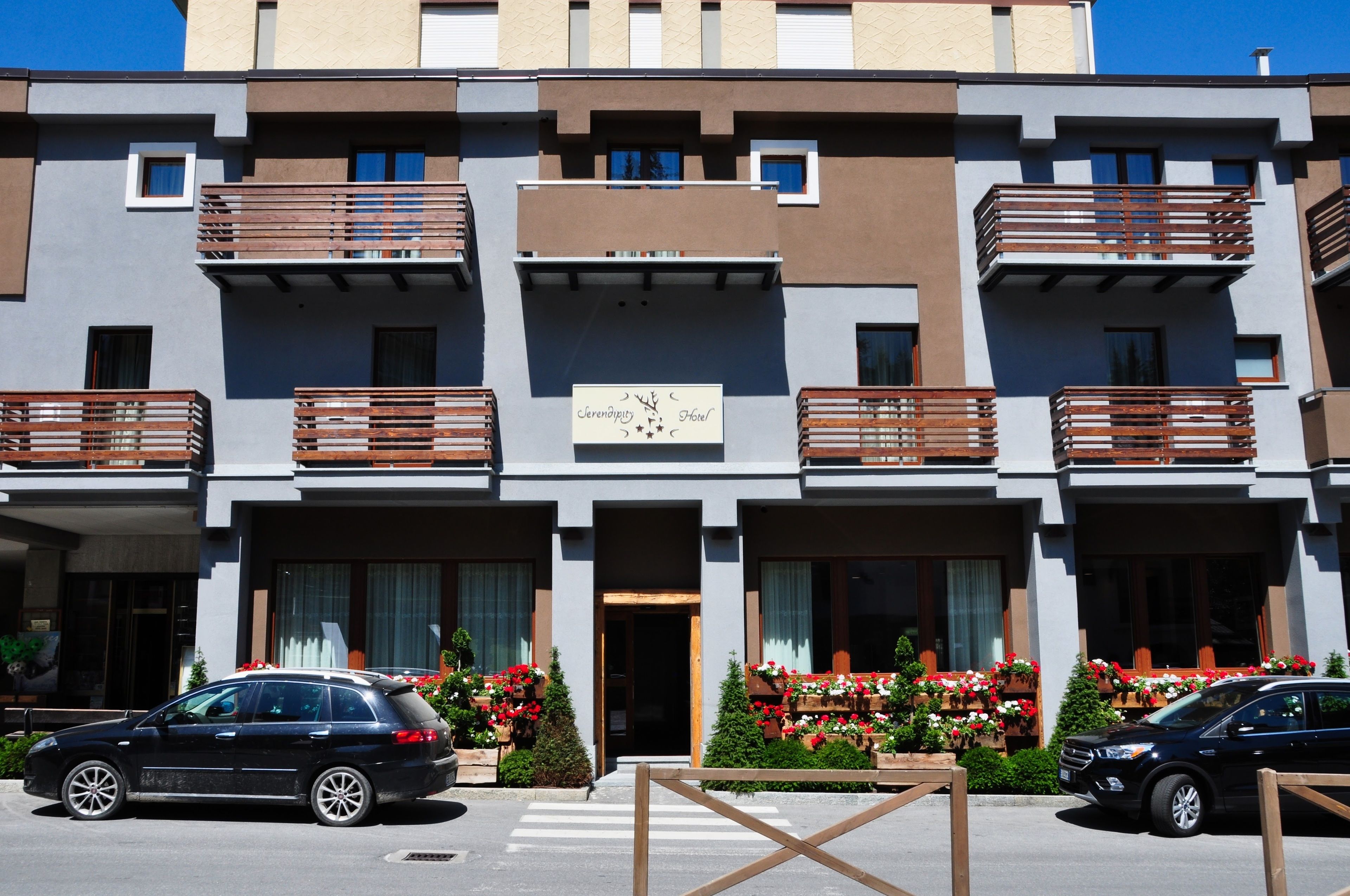 A modern hotel with a central entrance, balconies with wooden railings, and flower boxes with red and white flowers.