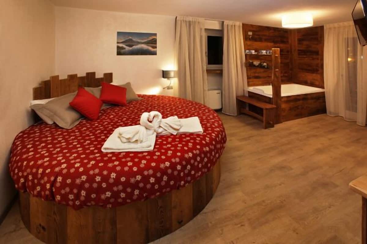 Un lit rond en bois, recouvert d’une courtepointe à motifs floraux rouges, avec deux oreillers rouges et une serviette blanche posée sur le lit.