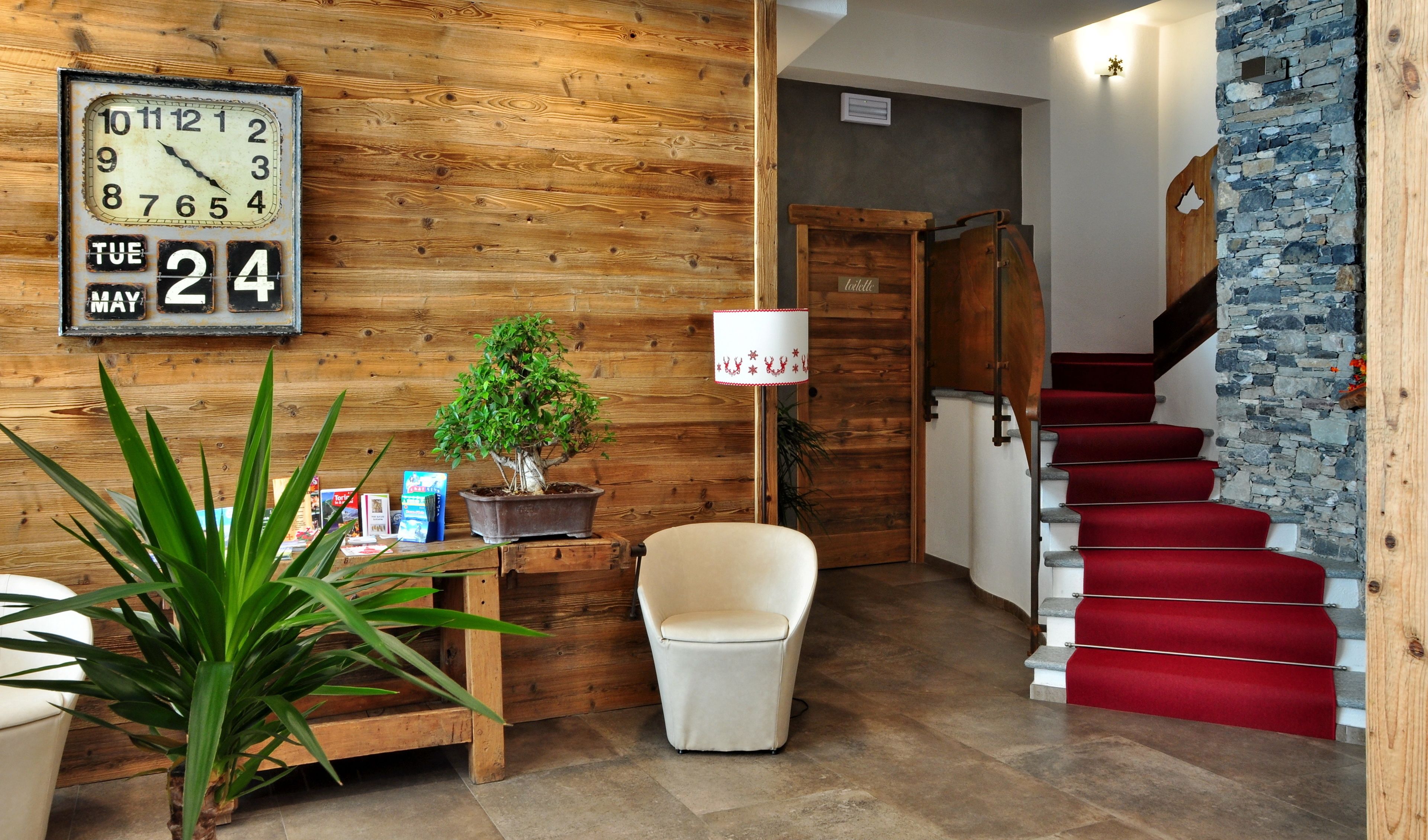 A wooden interior with a clock, a potted plant, a chair, and a red staircase.