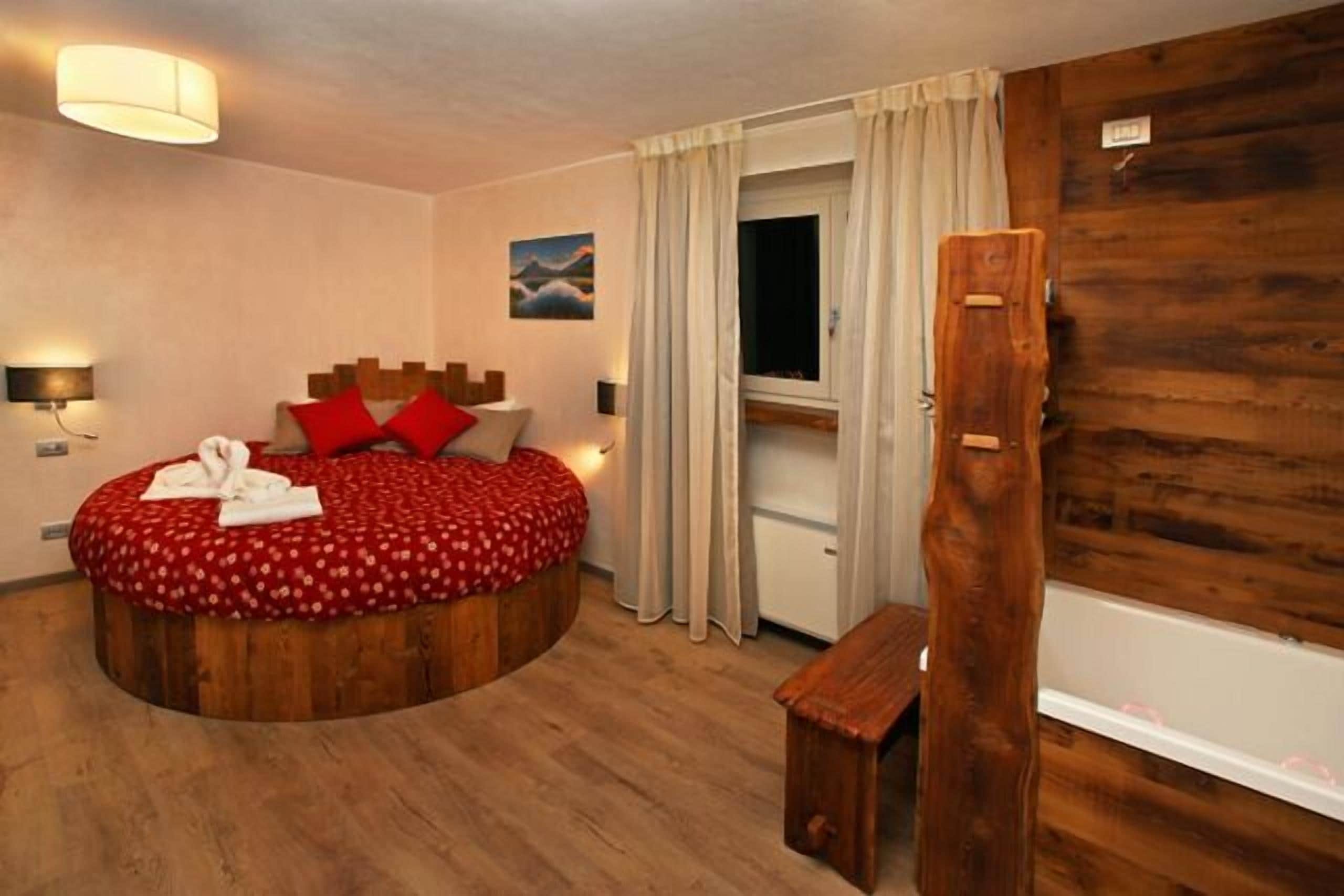 Ein rundes Holzbett mit roter Betttdecke und Kissen, eine Holzbank, eine Wandleuchte und ein Fenster mit Vorhängen.