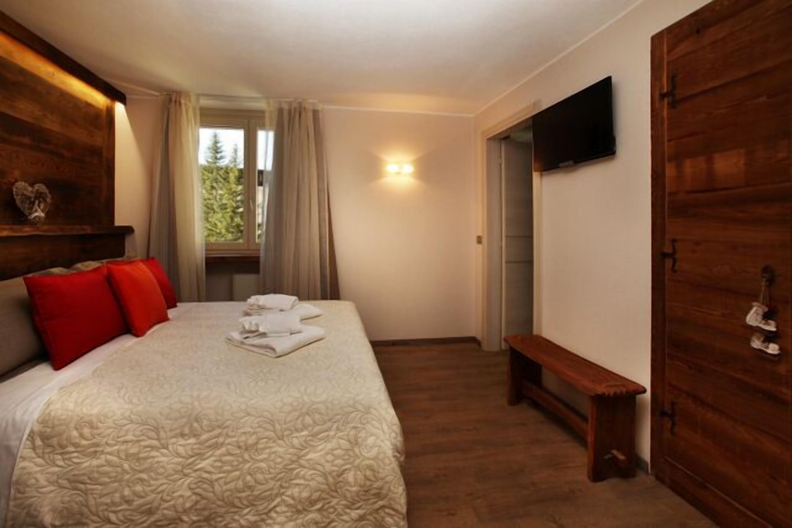 Un dormitorio con cama, un banco, televisor y ventana con cortinas.