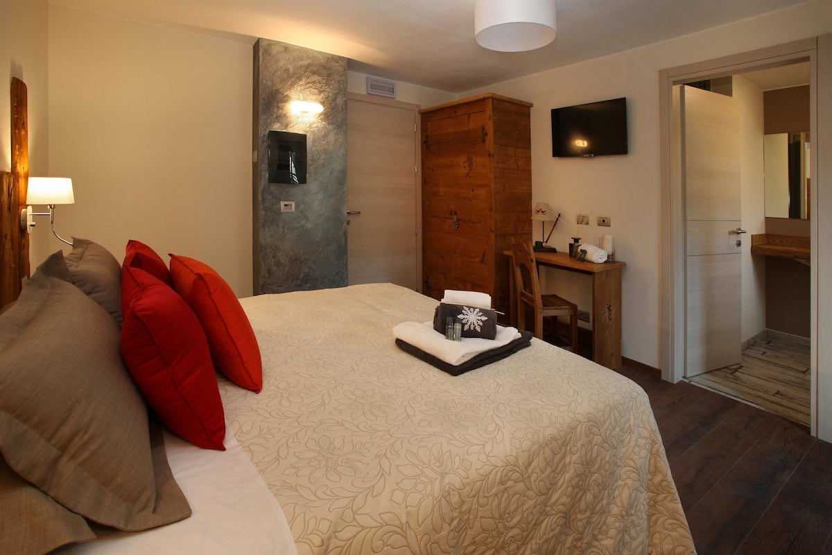 Une chambre d’hôtel avec un lit, une armoire en bois, un bureau avec une chaise, une télévision et une salle de bain visible à travers une porte ouverte.