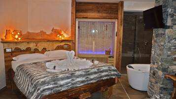 A bedroom with a wooden bed, a large window with curtains, a bathtub, and a television mounted on a stone wall.