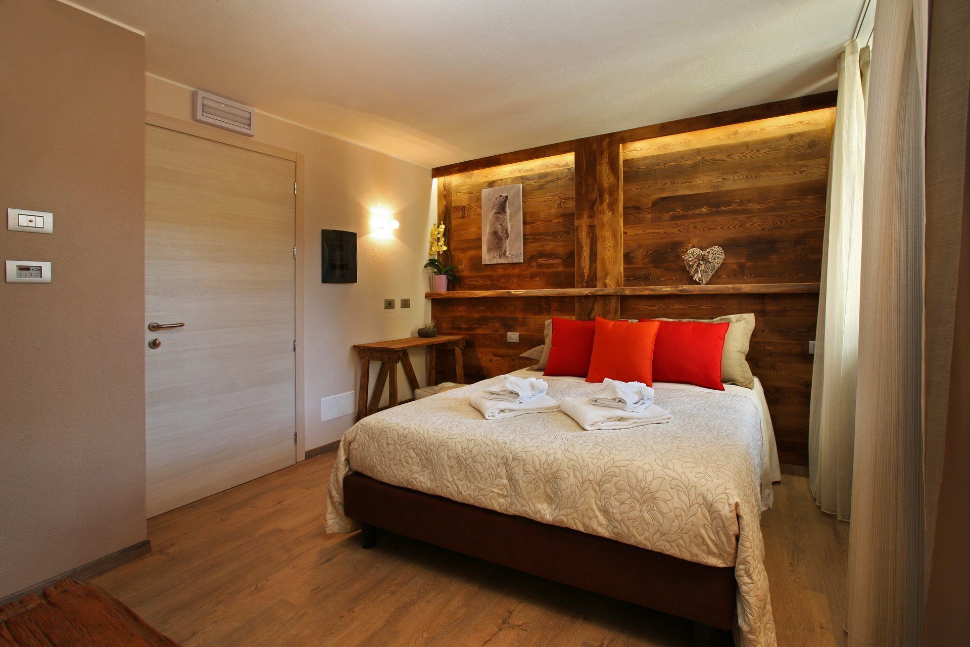 Een hotelkamer met een houten hoofdeinde, een bed met rode en beige kussens, een nachtkastje met een plant en een wandlamp.