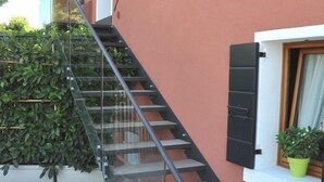 A modern staircase with glass railings and wooden steps, leading to a window with closed shutters and a potted plant on the windowsill. - Casa Rosanna (Meolo)
