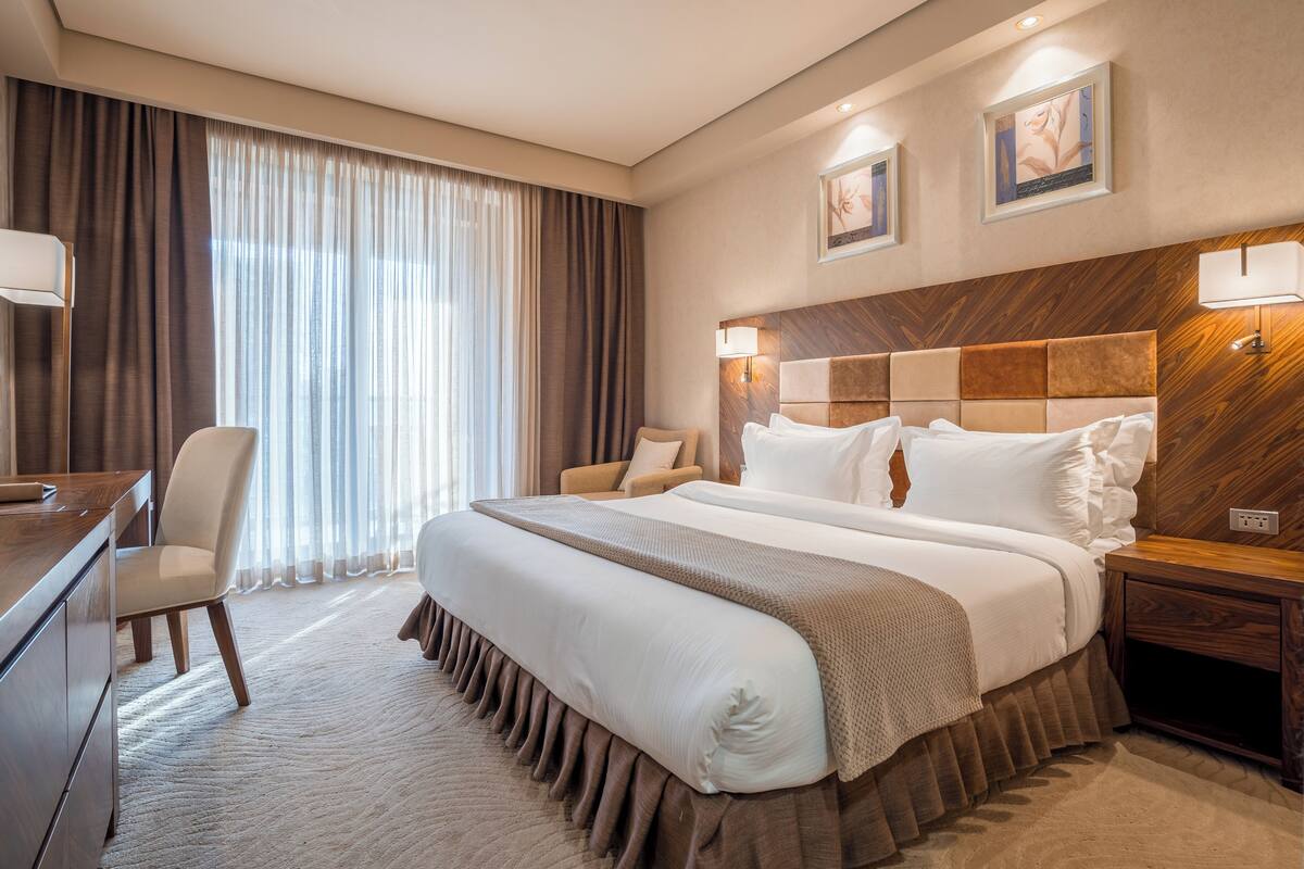 A hotel room with a large bed, a desk, a chair, and a nightstand with a lamp.