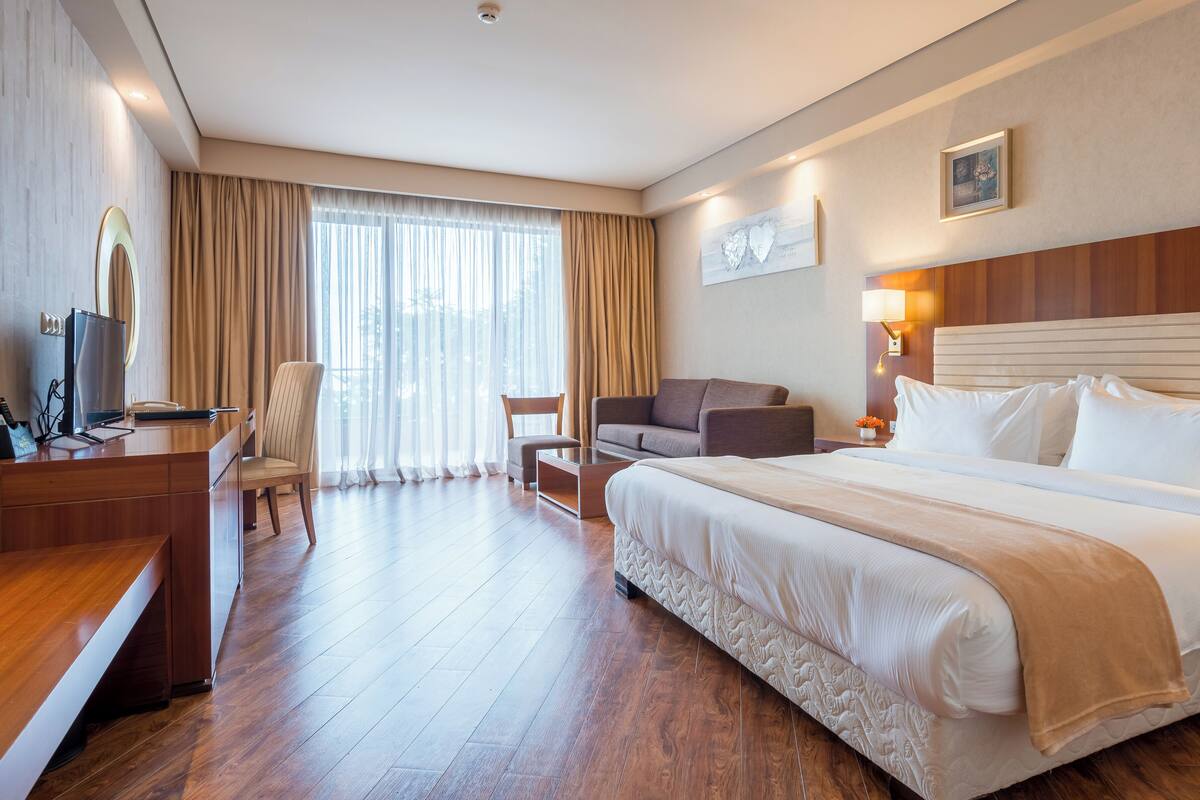 A hotel room with a large bed, a desk with a computer, a chair, a sofa, and a window with curtains.