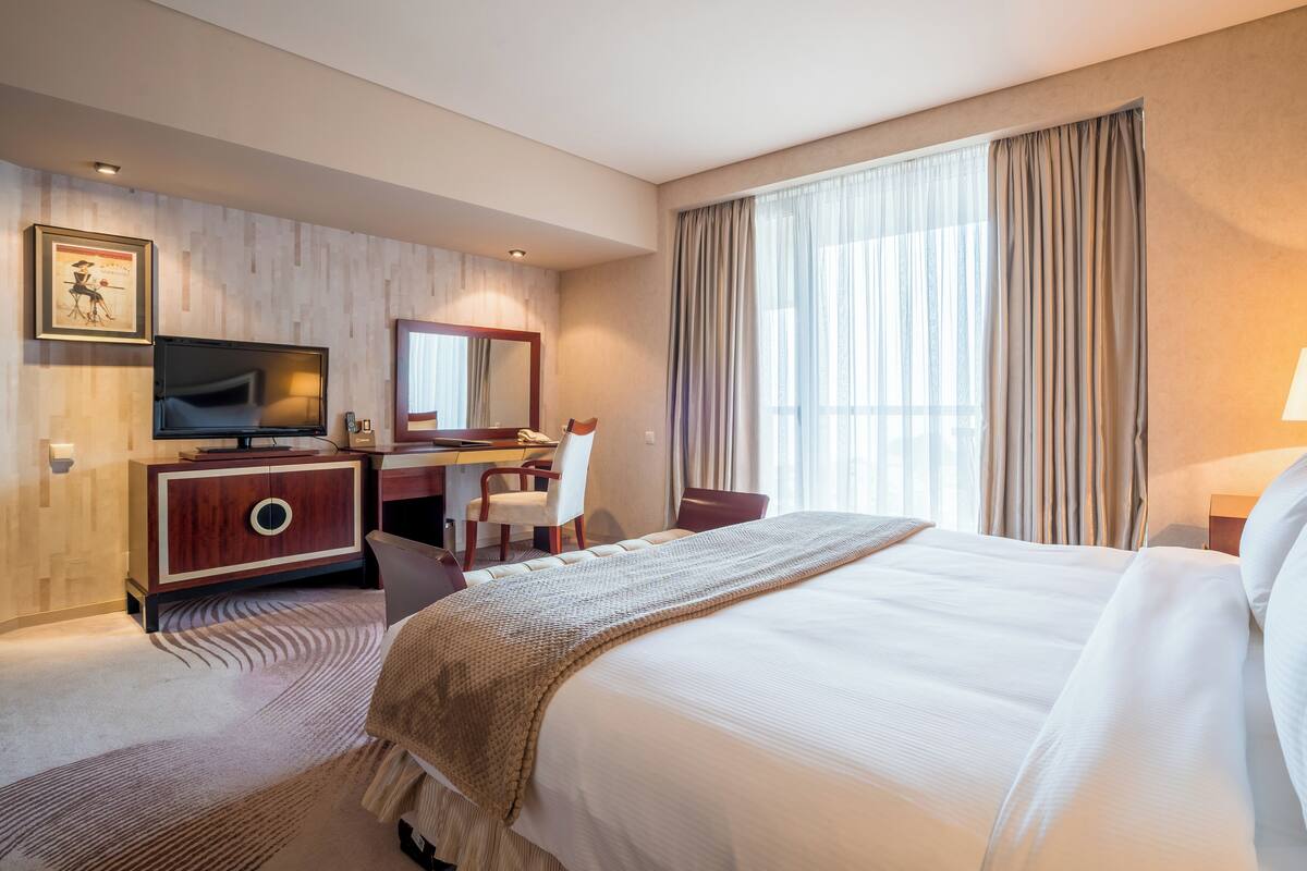 A hotel room with a large bed, a desk with a chair, a television, a mirror, and a window with curtains.