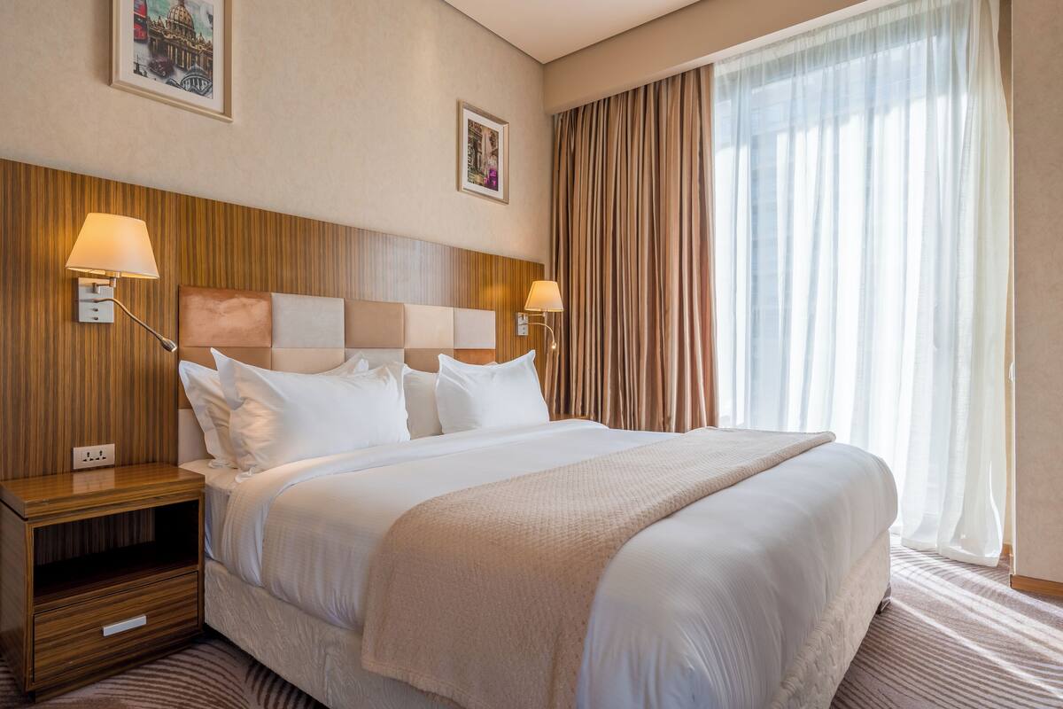 A hotel room with a large bed, two bedside tables, a wooden headboard, and a large window with curtains.