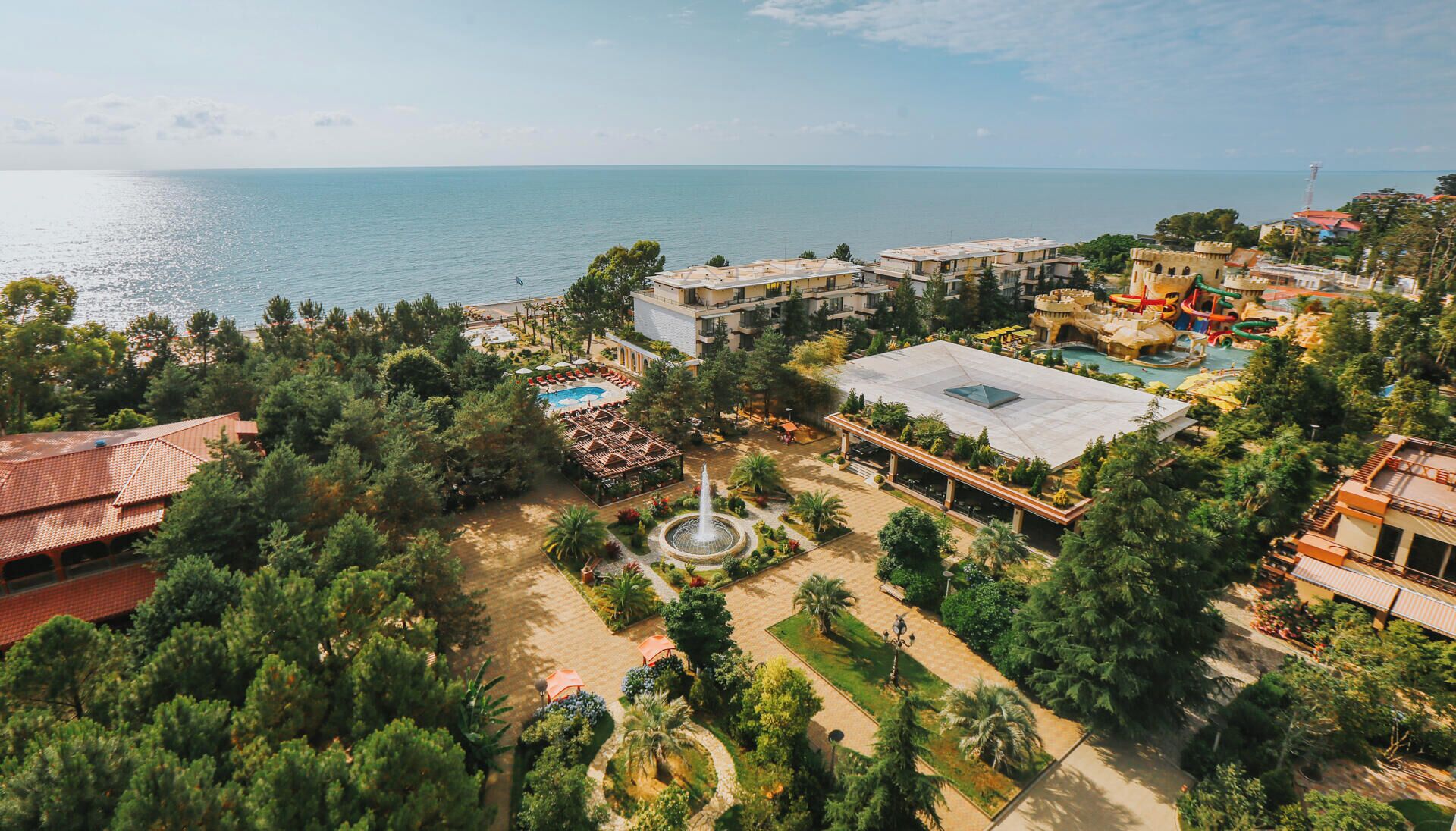 An aerial view of a resort with a central pool, a fountain, and a view of the sea.