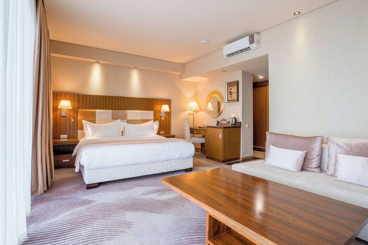 A hotel room with a large bed, a sofa, a wooden coffee table, a desk with a chair, and a wardrobe.