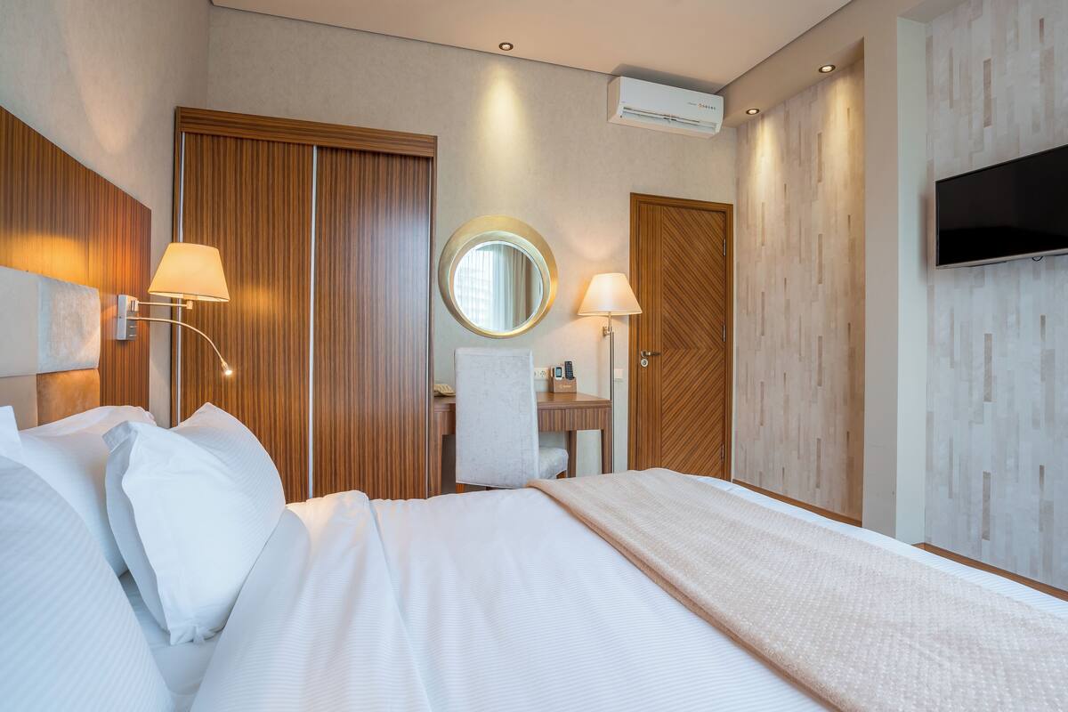 A hotel room with a bed, two wall-mounted lamps, a round mirror, a wooden wardrobe, a TV mounted on the wall, and an air conditioning unit.