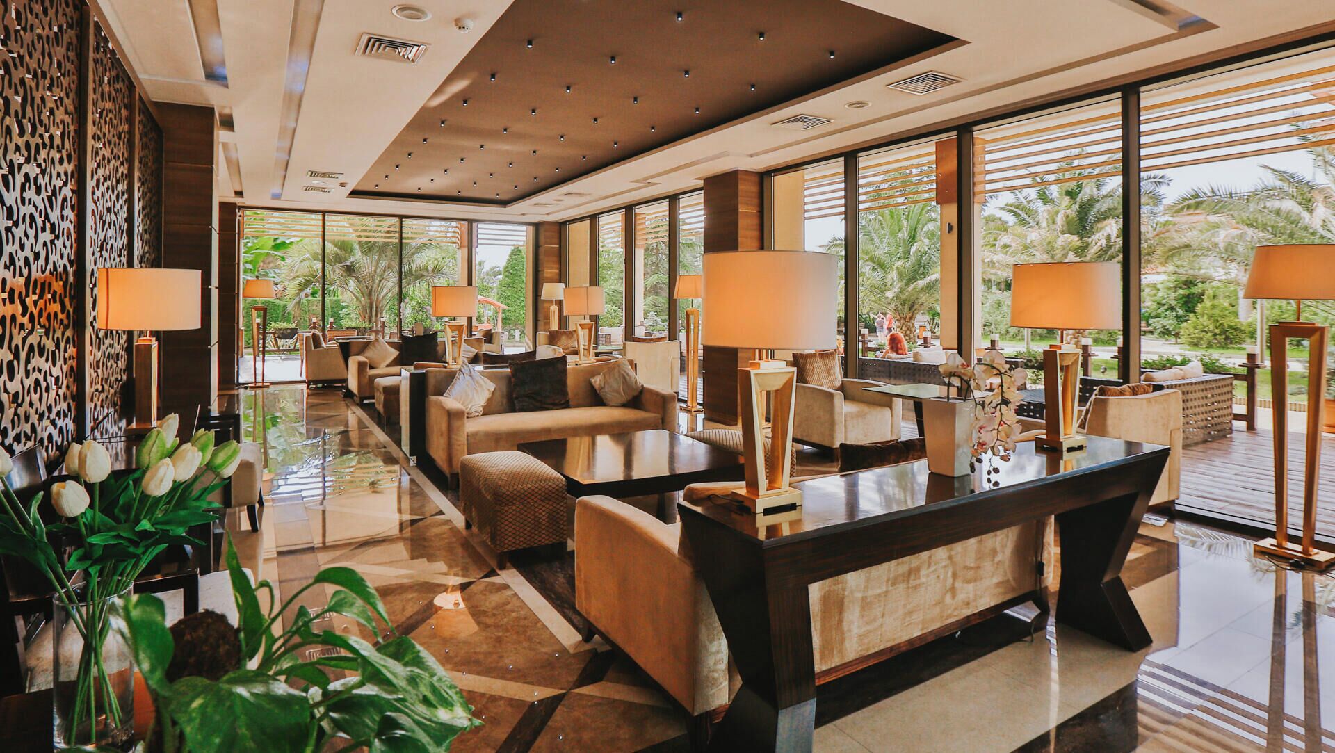 A modern hotel lobby with a reception desk, seating area, and large windows offering a view of palm trees.