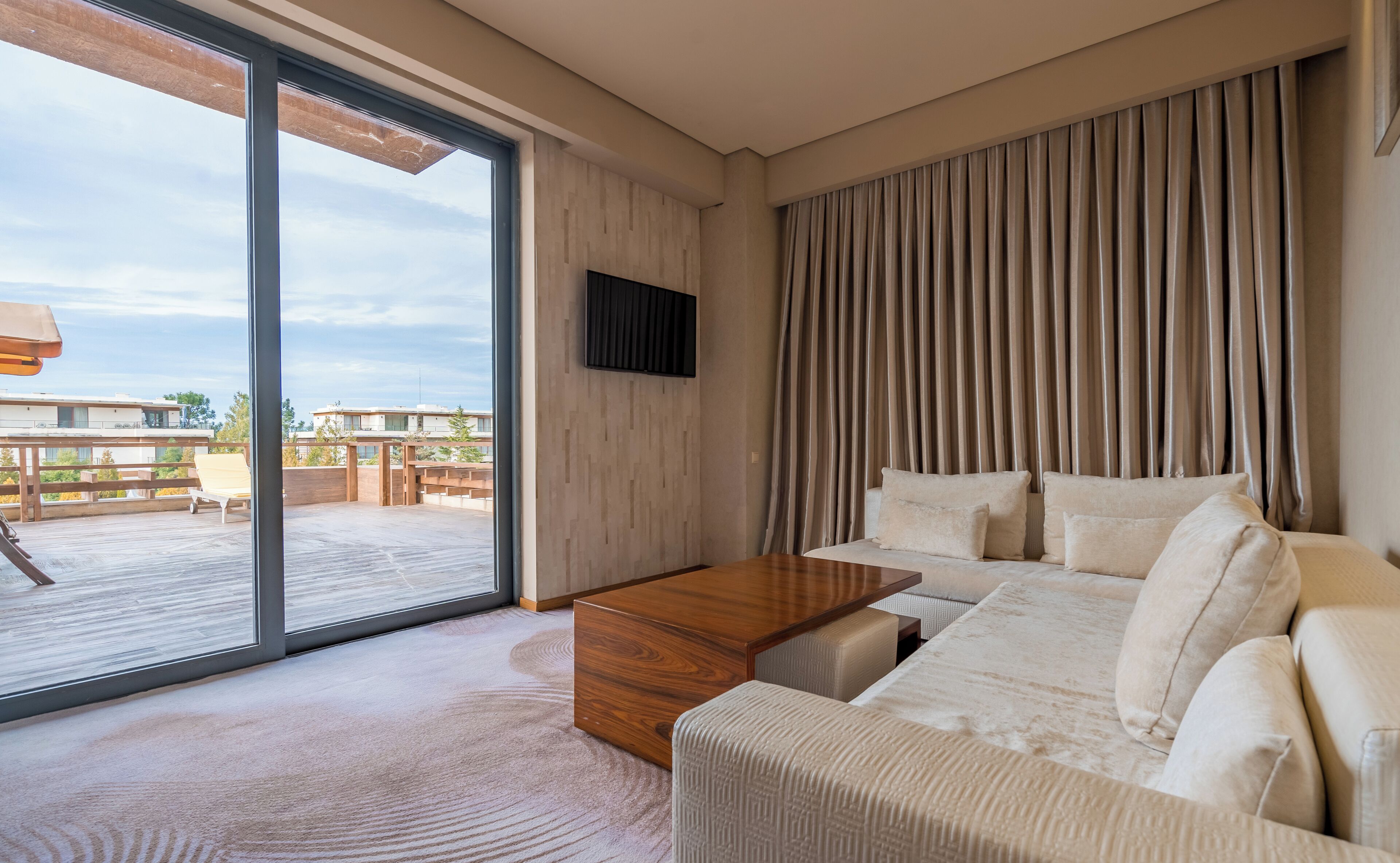 Una sala de estar moderna con un amplio ventanal corredero que da a una terraza exterior, una mesa de centro de madera, un sofá beige con cojines y un televisor de pantalla plana montado en la pared.