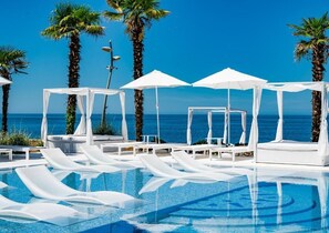 Zona junto a la piscina con sillones de descanso y sombrillas, palmeras y vistas al océano.