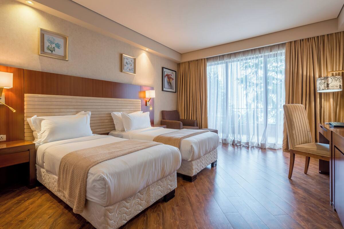 A hotel room with two beds, wooden headboard, bedside lamps, a desk, chair, and a large window with curtains.