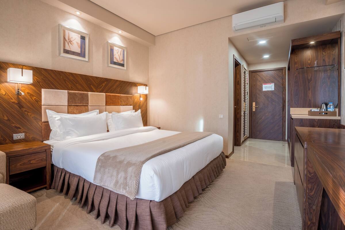 A hotel room with a large bed, wooden headboard, two wall-mounted lamps, a wooden wardrobe, and a small bedside table.