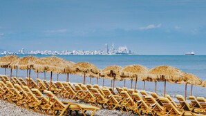 A beach with rows of yellow lounge chairs and thatched umbrellas, a city skyline in the background, and a ship on the water. - Dreamland Oasis Resort & Spa (Kobuleti)