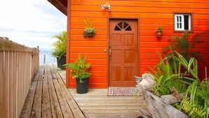 A wooden deck leading to a bright orange building with a brown door, potted plants, and a small window. - Orange House - Over the Water Rental (Bocas del Toro)