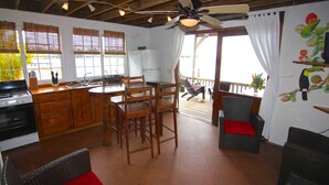 A kitchen with wooden cabinets, a dining table, and chairs. There is a ceiling fan, a refrigerator, and a window with blinds. - Orange House - Over the Water Rental (Bocas del Toro)