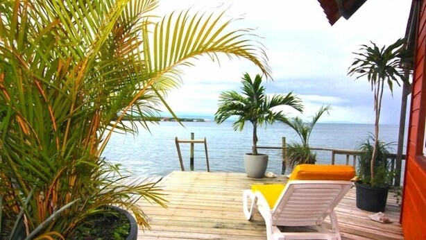 A wooden deck with a lounge chair and a table, overlooking a body of water with palm trees and a distant shoreline. - Orange House - Over the Water Rental (Bocas del Toro)