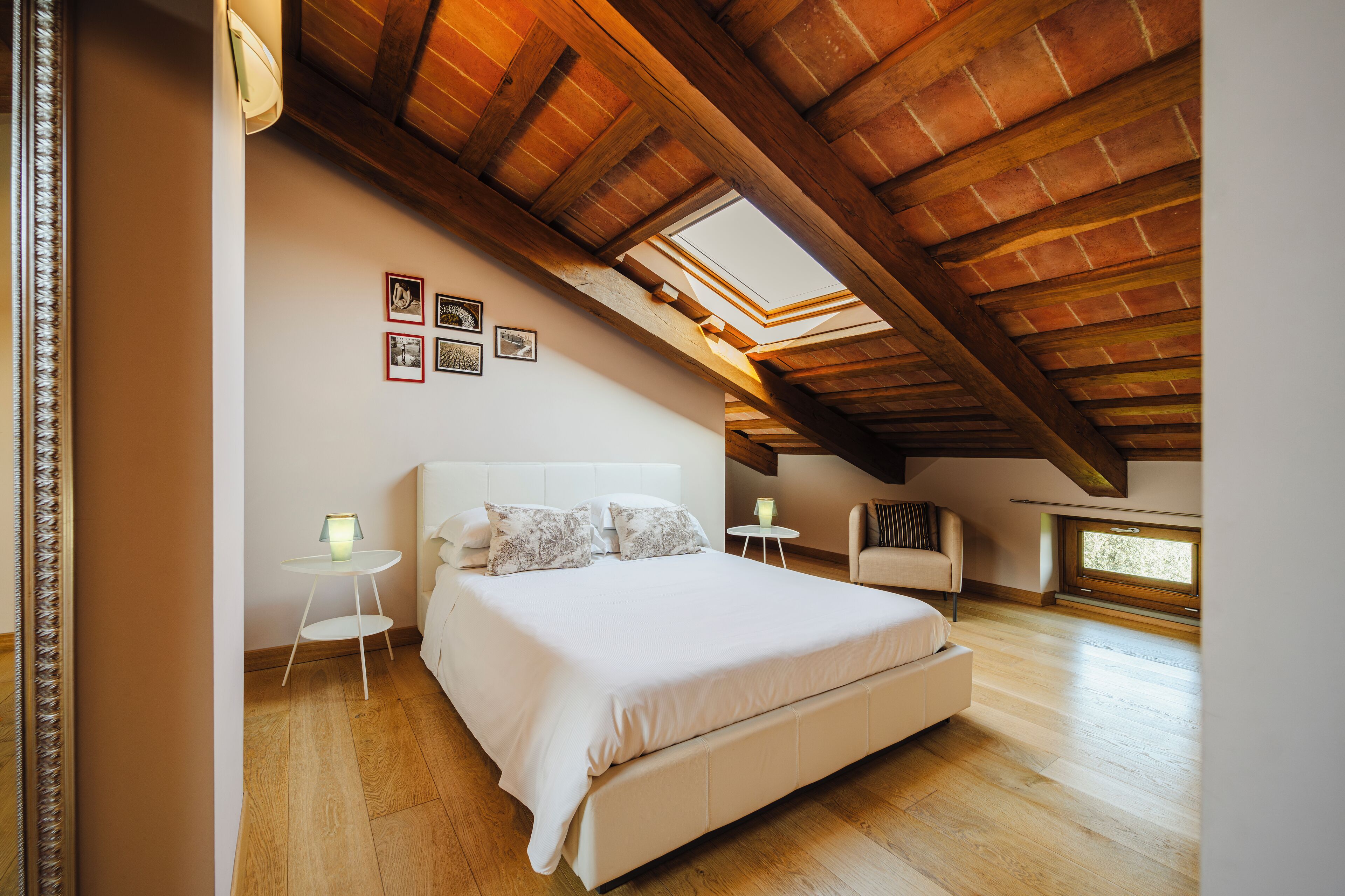 A bedroom with a large bed, a small table, and a chair. There are skylights and a window.