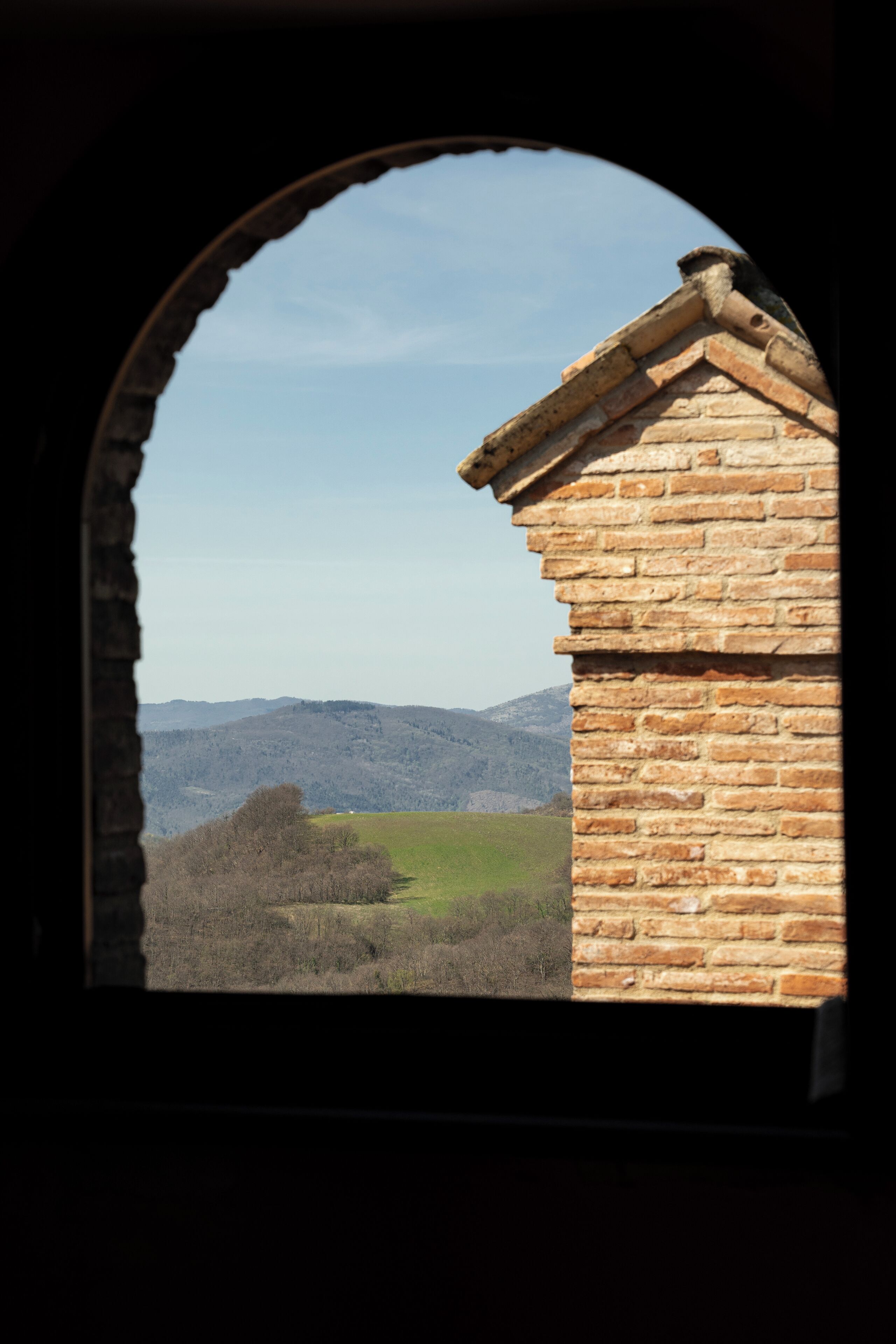 A view through an arched window of a brick building with a sloped roof, overlooking a hilly landscape with trees and distant mountains.