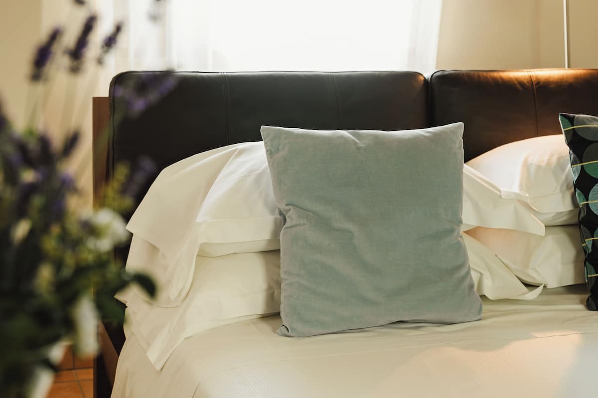 A neatly made bed with white linens and a single light blue pillow, a dark brown headboard, and a bedside lamp.