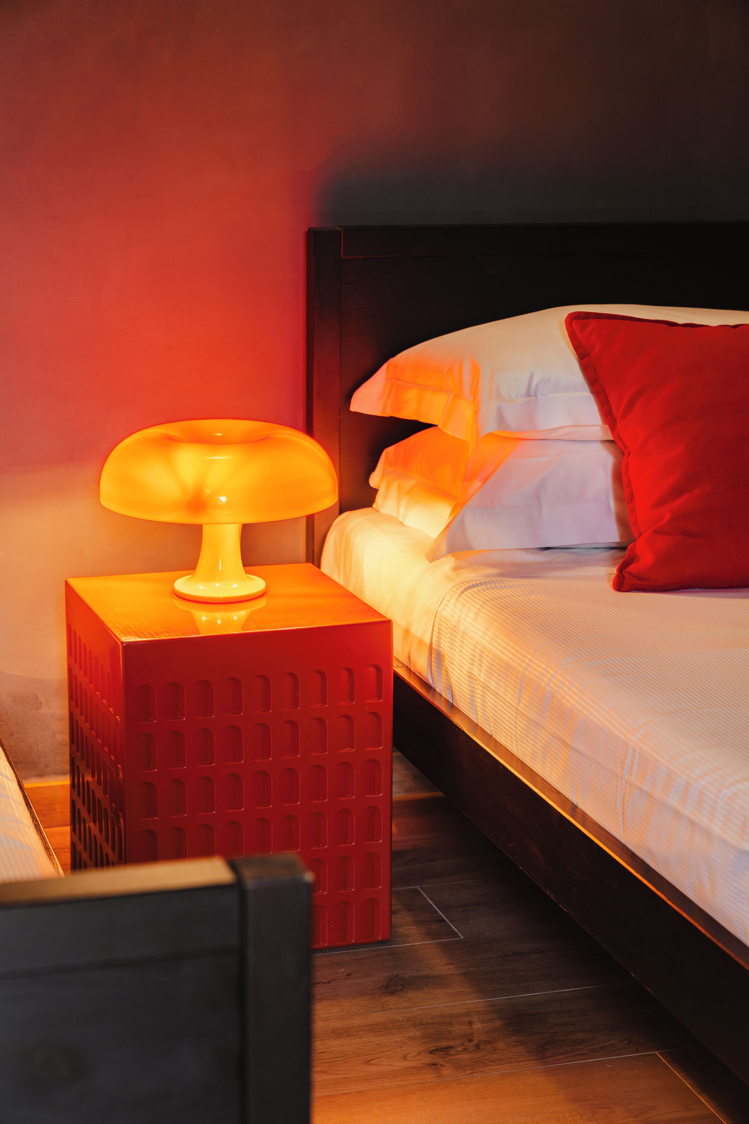 Un lit bien fait, avec une literie blanche et un oreiller rouge, une table de chevet rouge avec une lampe, et une tête de lit en bois foncé.