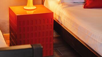 Un lit bien fait, avec une literie blanche et un oreiller rouge, une table de chevet rouge avec une lampe, et une tête de lit en bois foncé.