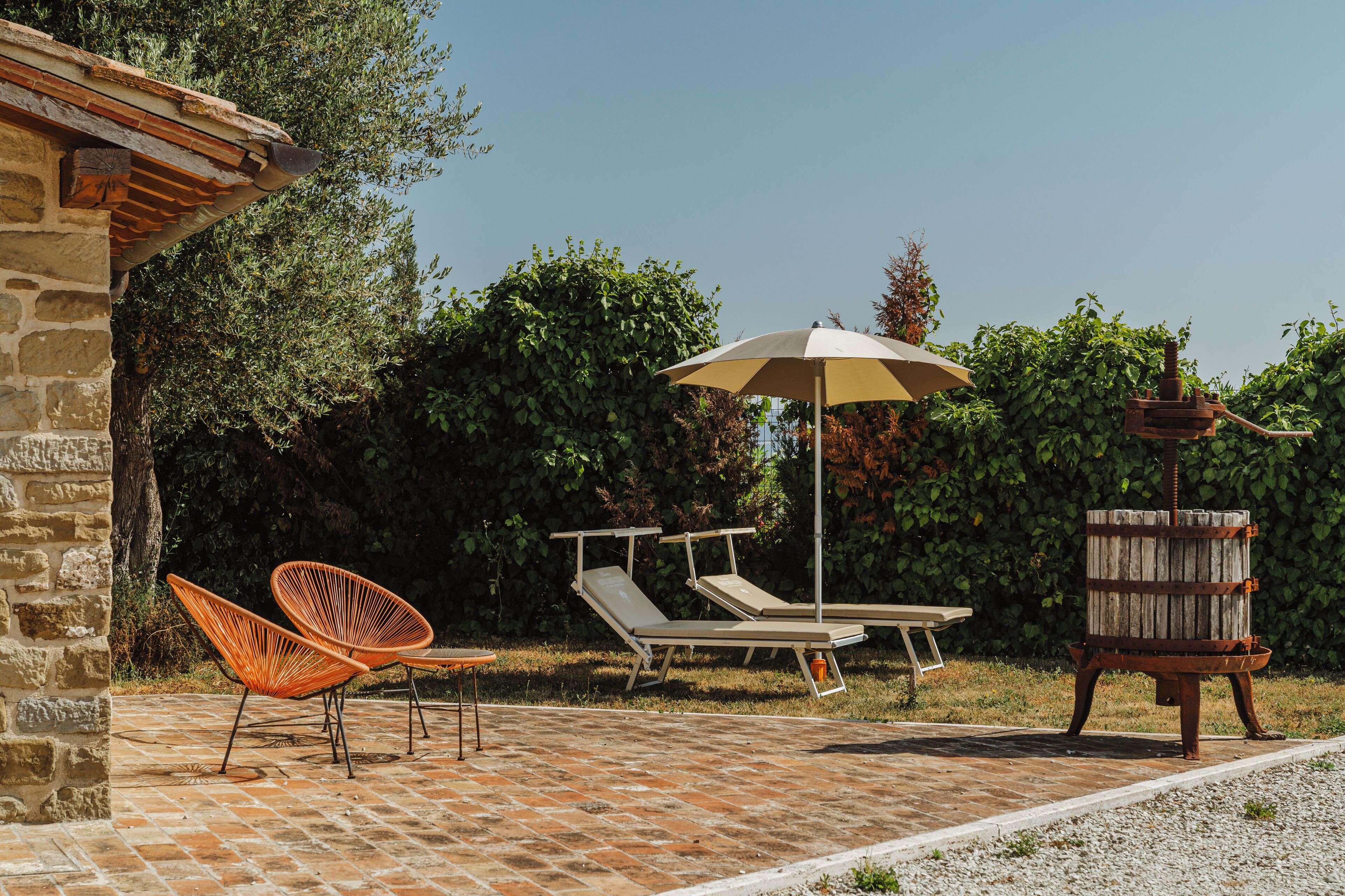 An outdoor seating area with lounge chairs and a table, surrounded by greenery and a stone wall.