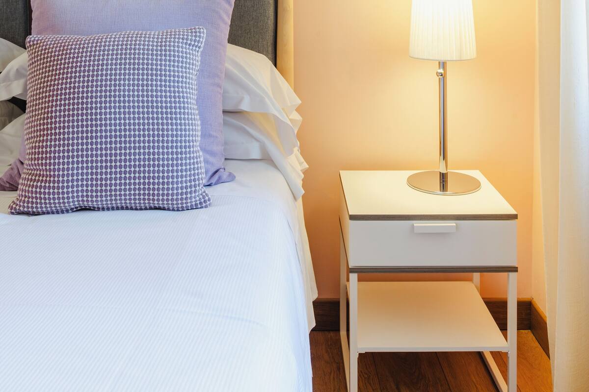 A neatly made bed with a grey headboard, a white bedside table with a lamp, and a patterned pillow.
